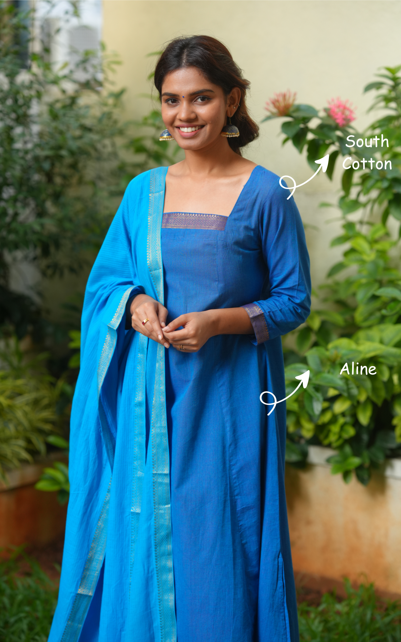 Woman wearing a blue traditional outfit with a dupatta, standing outdoors with plants in the background.